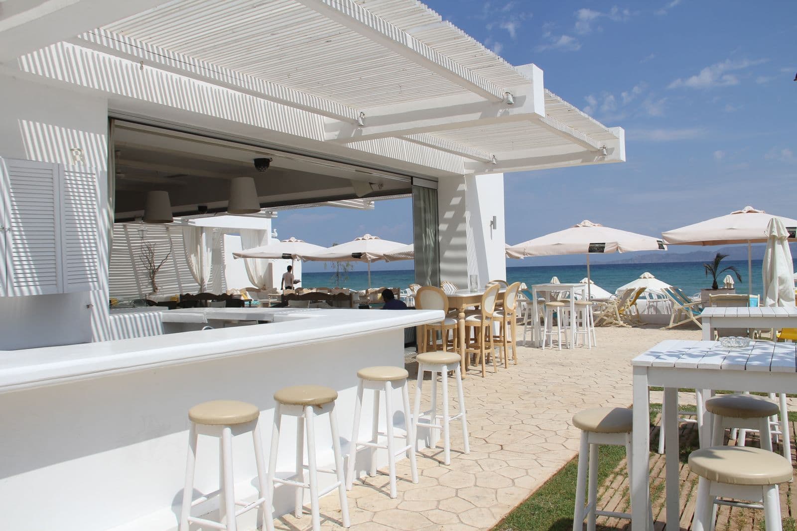 Le comptoir blanc du bar sous la pergola, tabourets et tables dressées face à la mer.