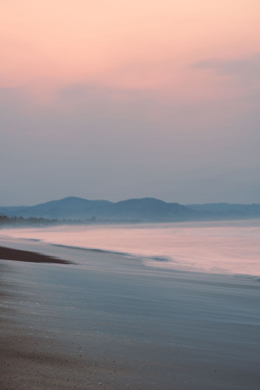 La plage à la tombée du jour, ciel rose pâle, mer étale et collines à l'horizon.